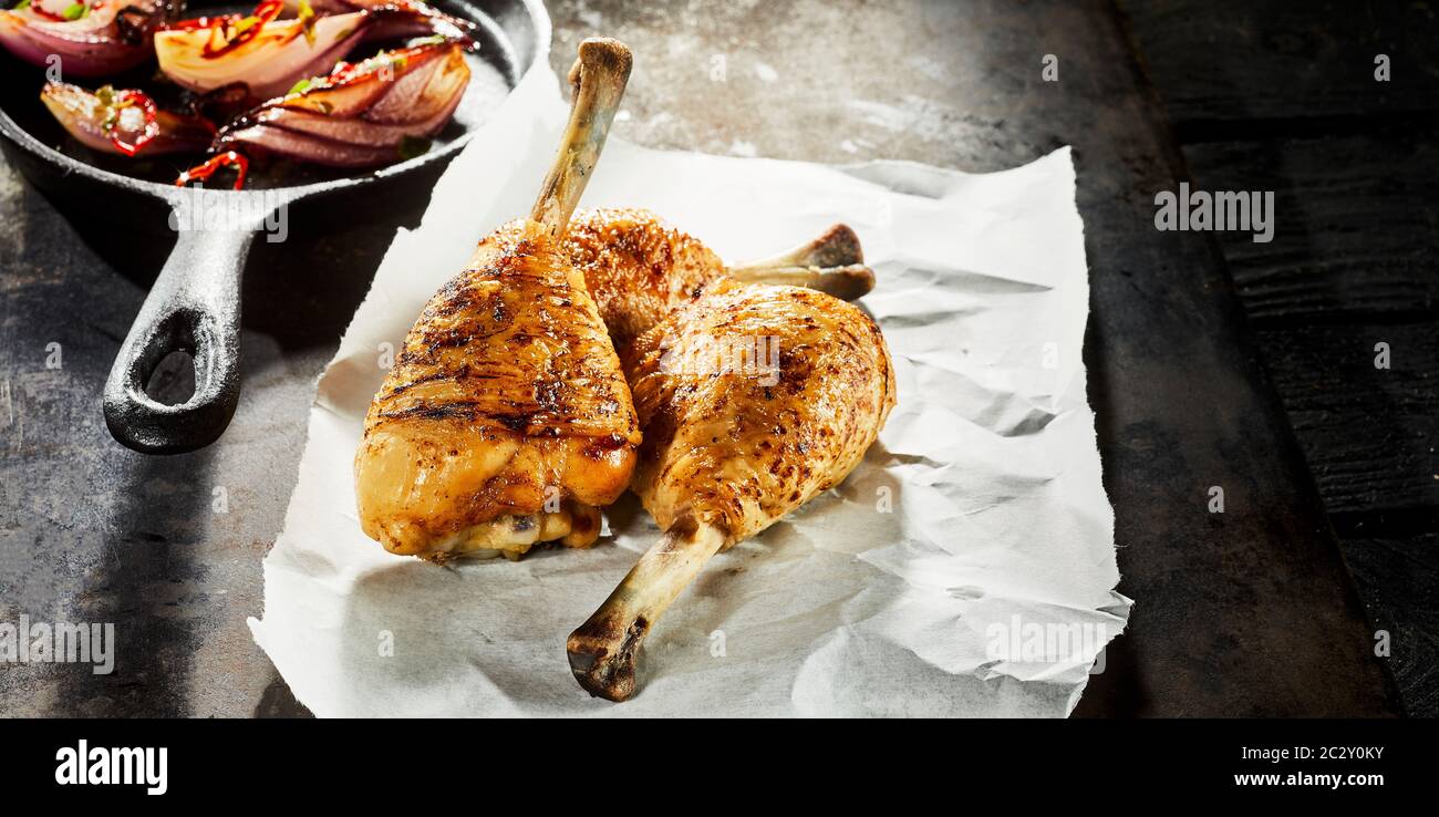 Three grilled spicy seasoned chicken drumsticks served on crumpled white paper with sauteed or roasted vegetables in a skillet Stock Photo