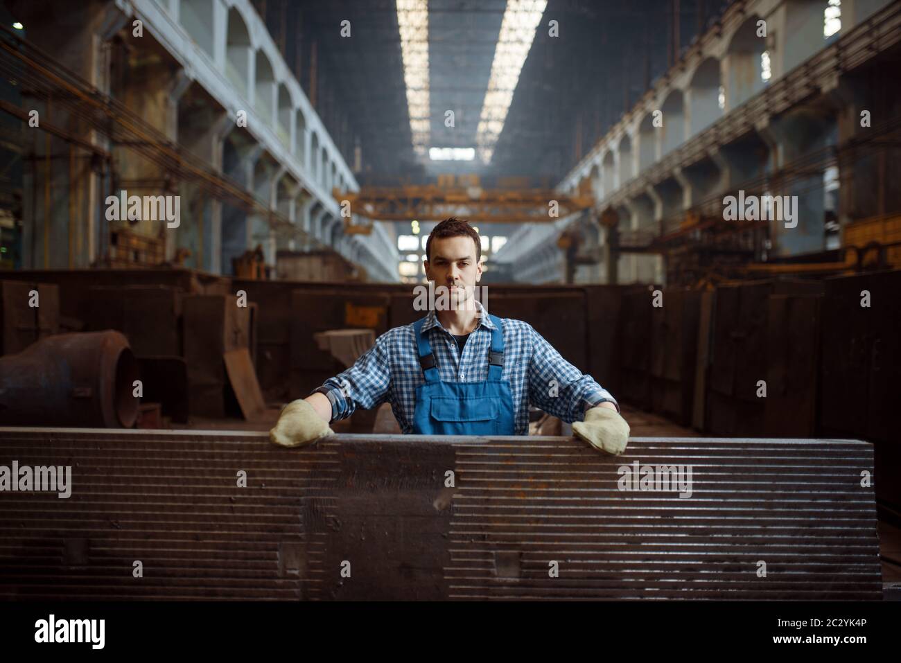 Male worker in uniform and gloves at stack of metal workpieces on factory. Metalworking industry, industrial manufacturing of steel products Stock Photo