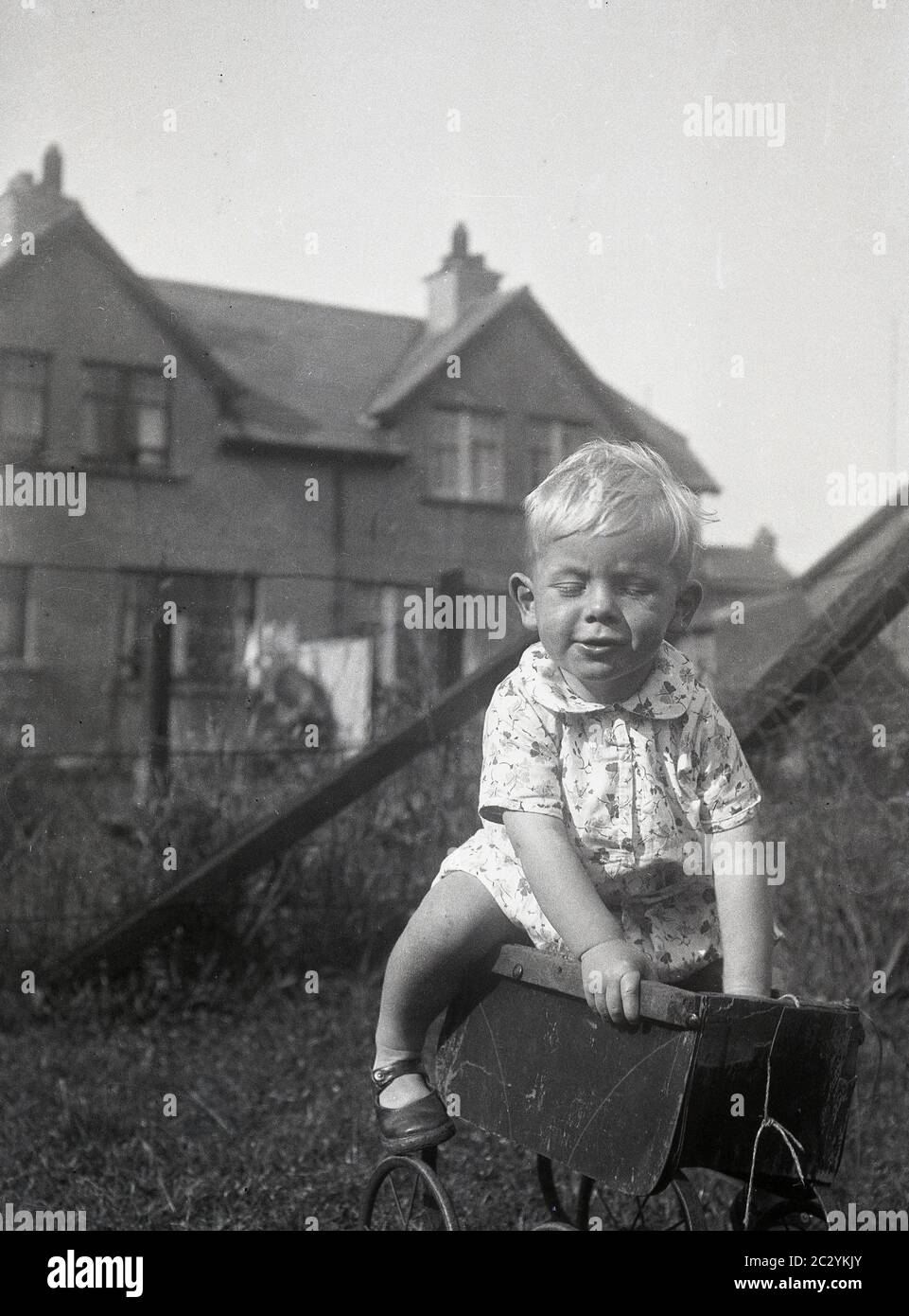 circa 1930s, historical, daytime and a little boy playing outside in a garden, climbing on top of an old metal wheeled child's toy or doll's pram of the era, which has lost its hood and handle, An interesting object to try and climb on and the little child shows good balance! Stock Photo