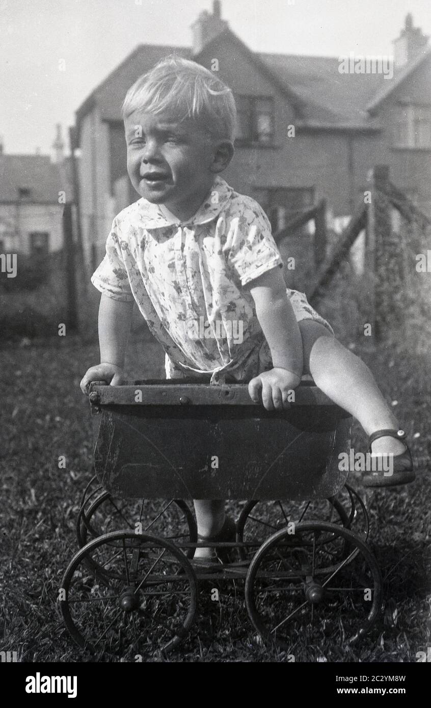 circa1930s, historical, daytime and a little boy playing outside in a garden, climbing on top of an old metal wheeled child's toy or doll's pram of the era, which has lost its hood and handle, An interesting object to try and climb on and the little child shows good balance! Stock Photo