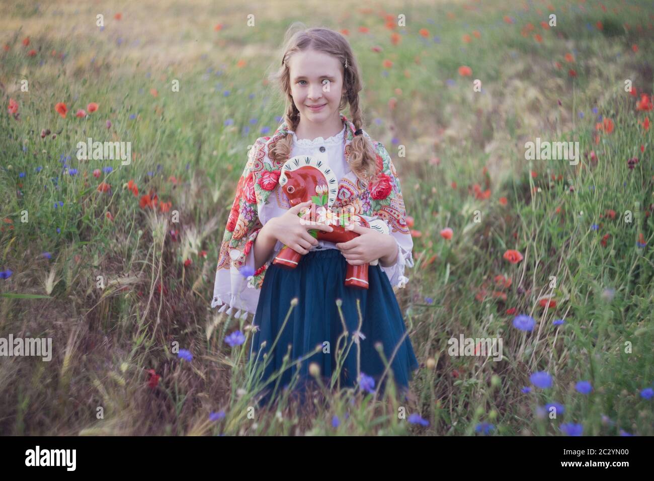 Portrait of a girl in a field with wild flowers. On the girl is a shawl with folk patterns. In her hands is a wooden horse painted in the style of lub Stock Photo