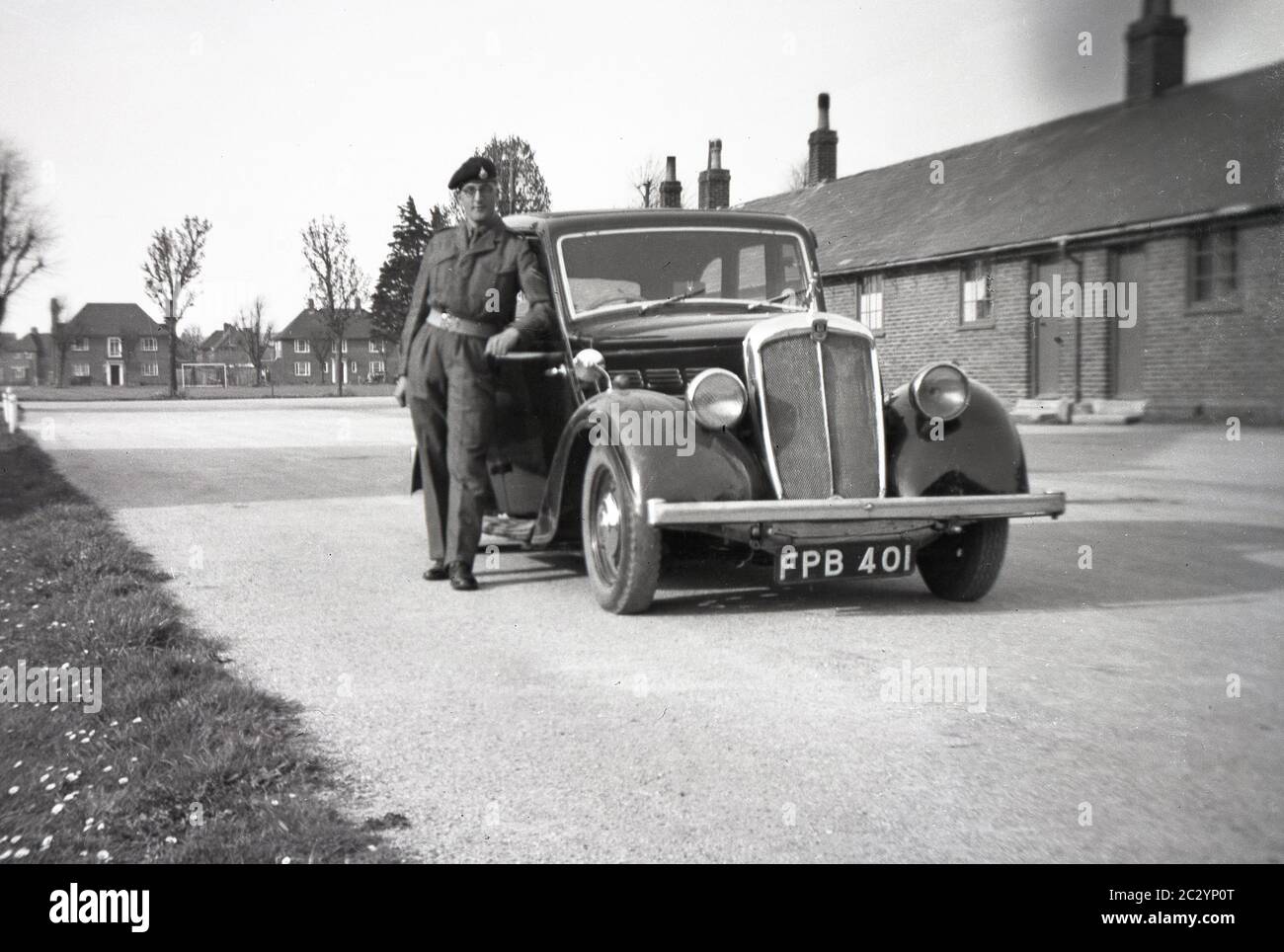 1940s, historical, a young soldier/driver in his uniform standing at an army base on a flat gravel area beside his motorcar, England, UK. The housing and accommodation of the senior Army Officers can be seen in the background. Stock Photo