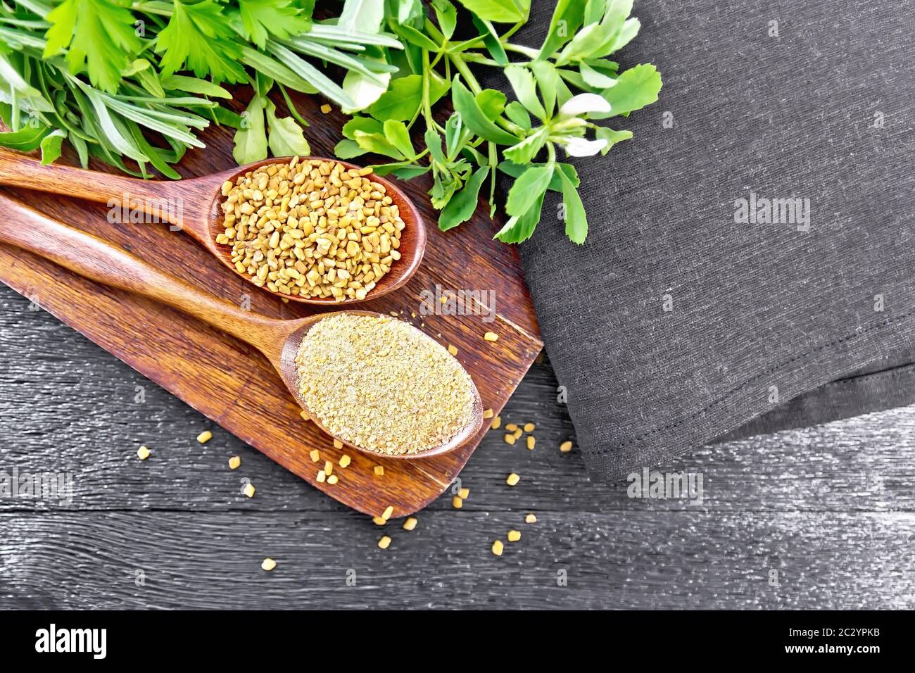 Fenugreek seeds and ground spice in two spoons on brown plate with herbs, a napkin on wooden board background from above Stock Photo