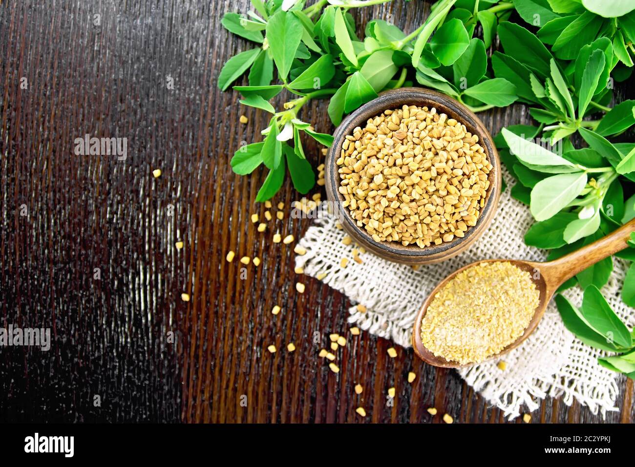 Ground fenugreek in a spoon and whole in a bowl on burlap with green leaves against dark wooden board on top Stock Photo