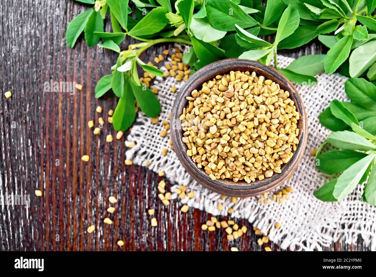 Fenugreek seeds in a bowl on burlap with green leaves on a wooden board background from above Stock Photo