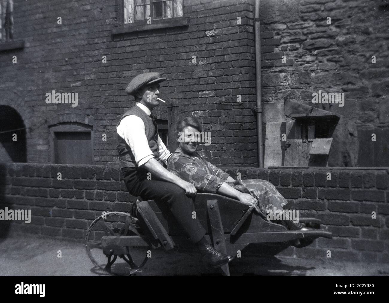 Man in a cloth cap and waistcoat and a cigarette in his mouth, sitting with a young lady on an old wooden framed metal wheelbarrow outside a door covered victorian brick building, common in the industrial areas of Northern England, Sheffield, circa, mid 1930s-1940s. Stock Photo