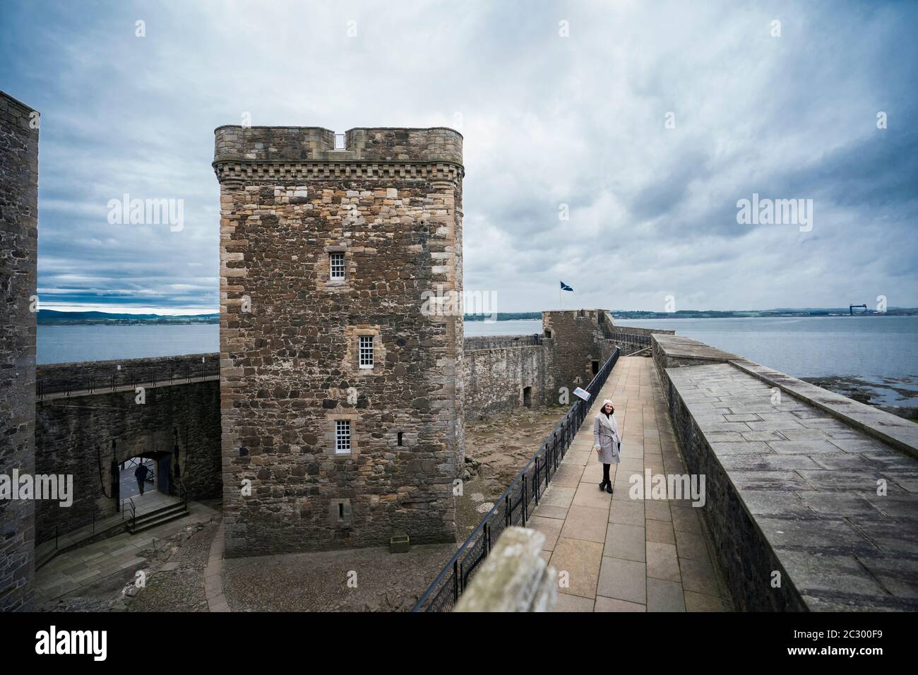 A beautiful woman wearing a warm white fur-collared coat and woolen hat walks down the balcony overlooking Blackness Castle shaped like a ship while v Stock Photo