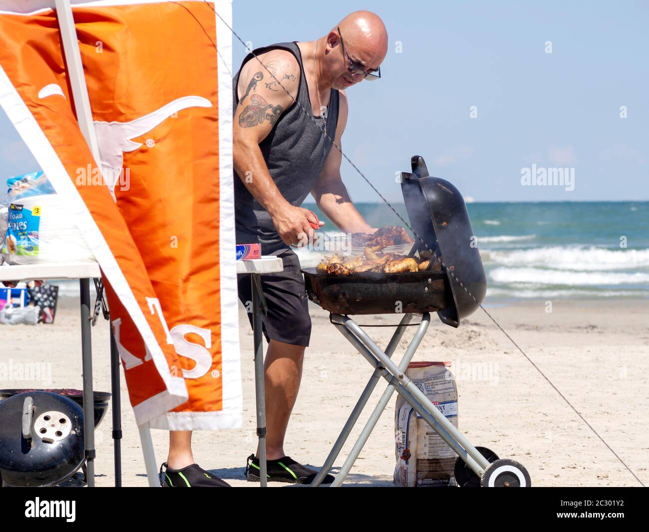 A bald man in sunglasses cooks chicken at a hot, smoky grill on a sunny day at the beach in Port Aransas, Texas USA. Stock Photo