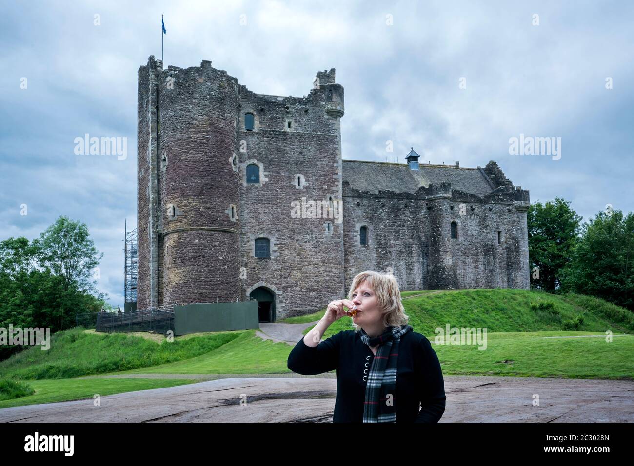Blond tourist in her 50's drinks a dram of whisky outdoors in front of Doune castle, Stirling, Scotland, UK, Europe Stock Photo