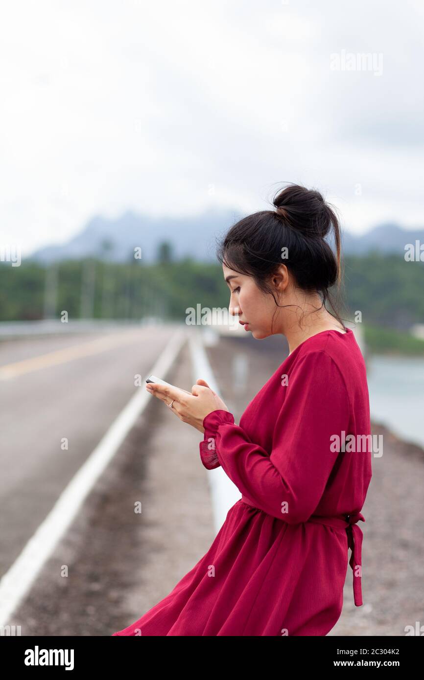 Portrait of beautiful young asian woman with Carmen red dress sitting on guard rail and using her smartphone Stock Photo