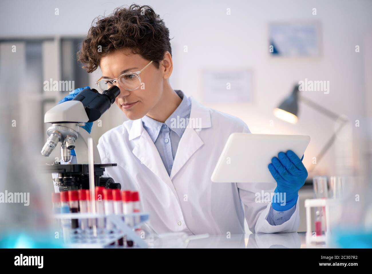 Concentrated laboratory technician using data on tablet while analyzing sample via microscope Stock Photo