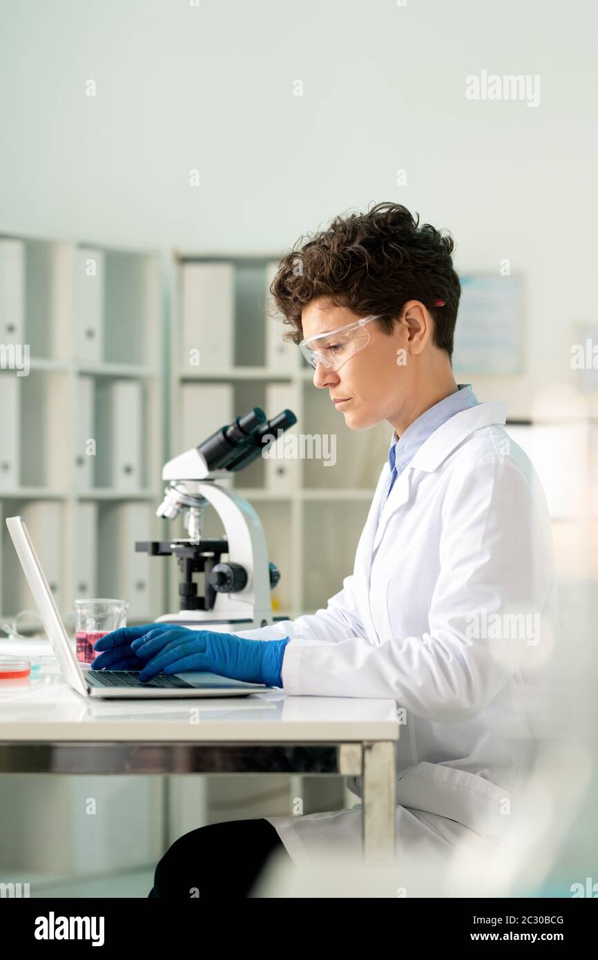 Concentrated virologist in protective goggles and gloves typing on laptop while working on coronavirus research in laboratory Stock Photo