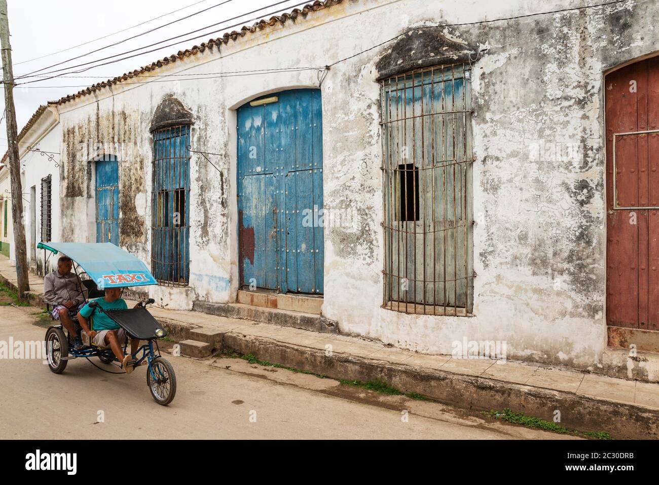 Bicycle taxi driving past decayed facades of crumbling plaster and weathered wooden doors, Remedios, Cuba Stock Photo