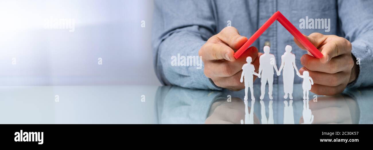 Close-up Of A Businesswoman's Hand Protecting Family Figures With Red Roof On Reflective Desk Stock Photo
