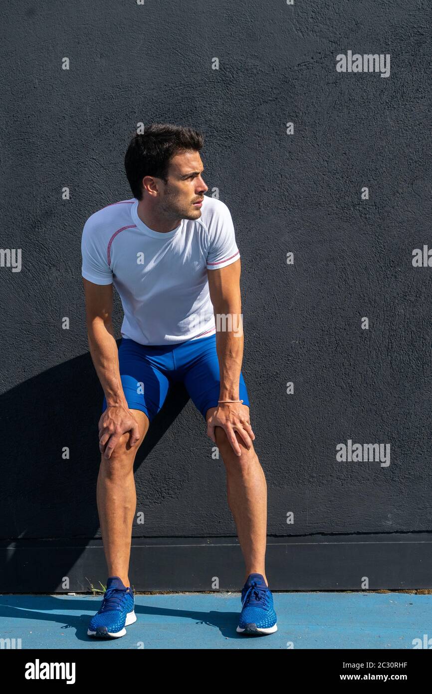 Young running man in blue pants and white shirt resting and recovering on black wall in full sun. Motivation concept, career Stock Photo