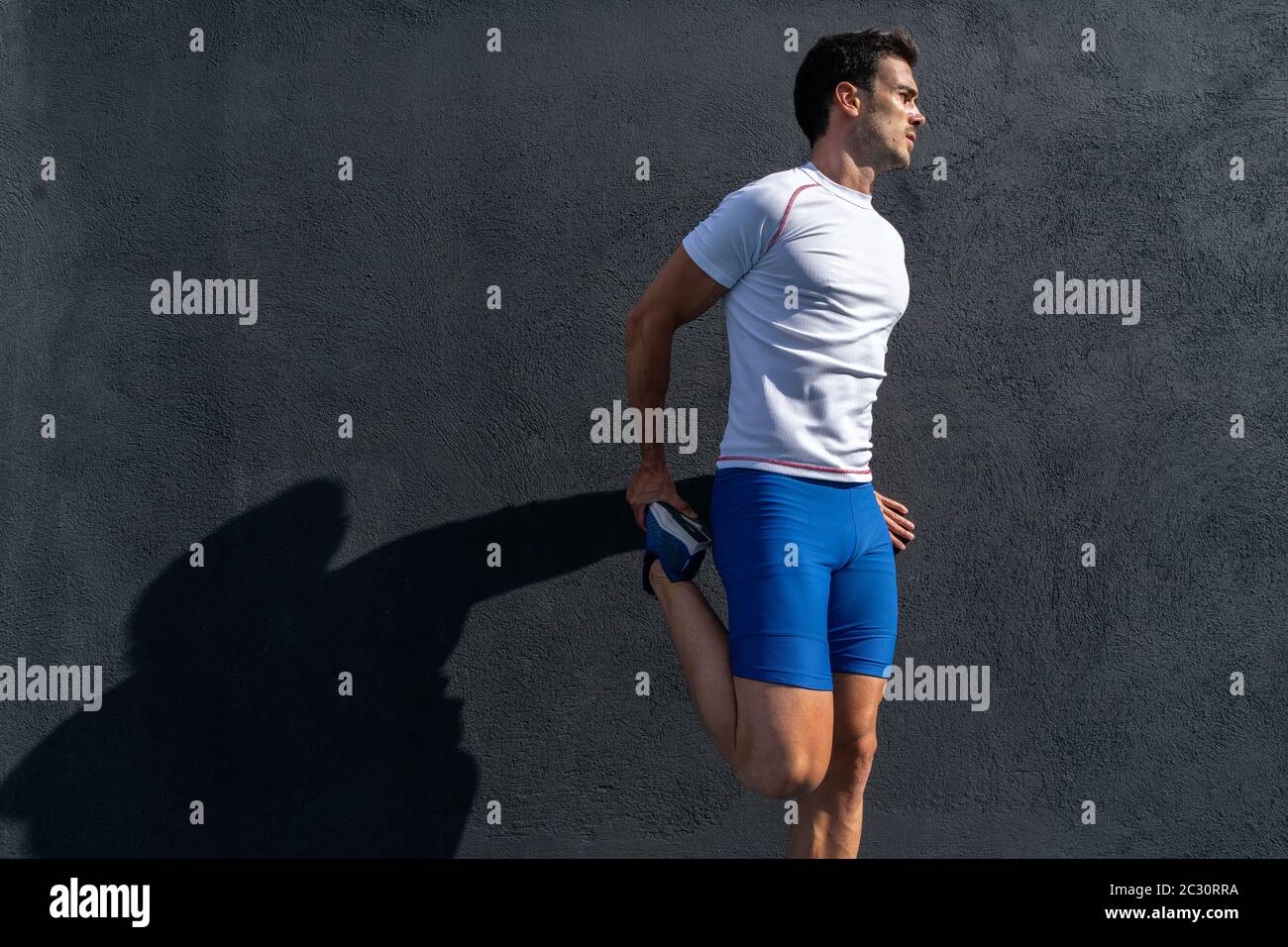 Young man running in blue pants and white shirt resting and recovering on black wall in full sun, doing leg stretches. Motivation concept, career Stock Photo