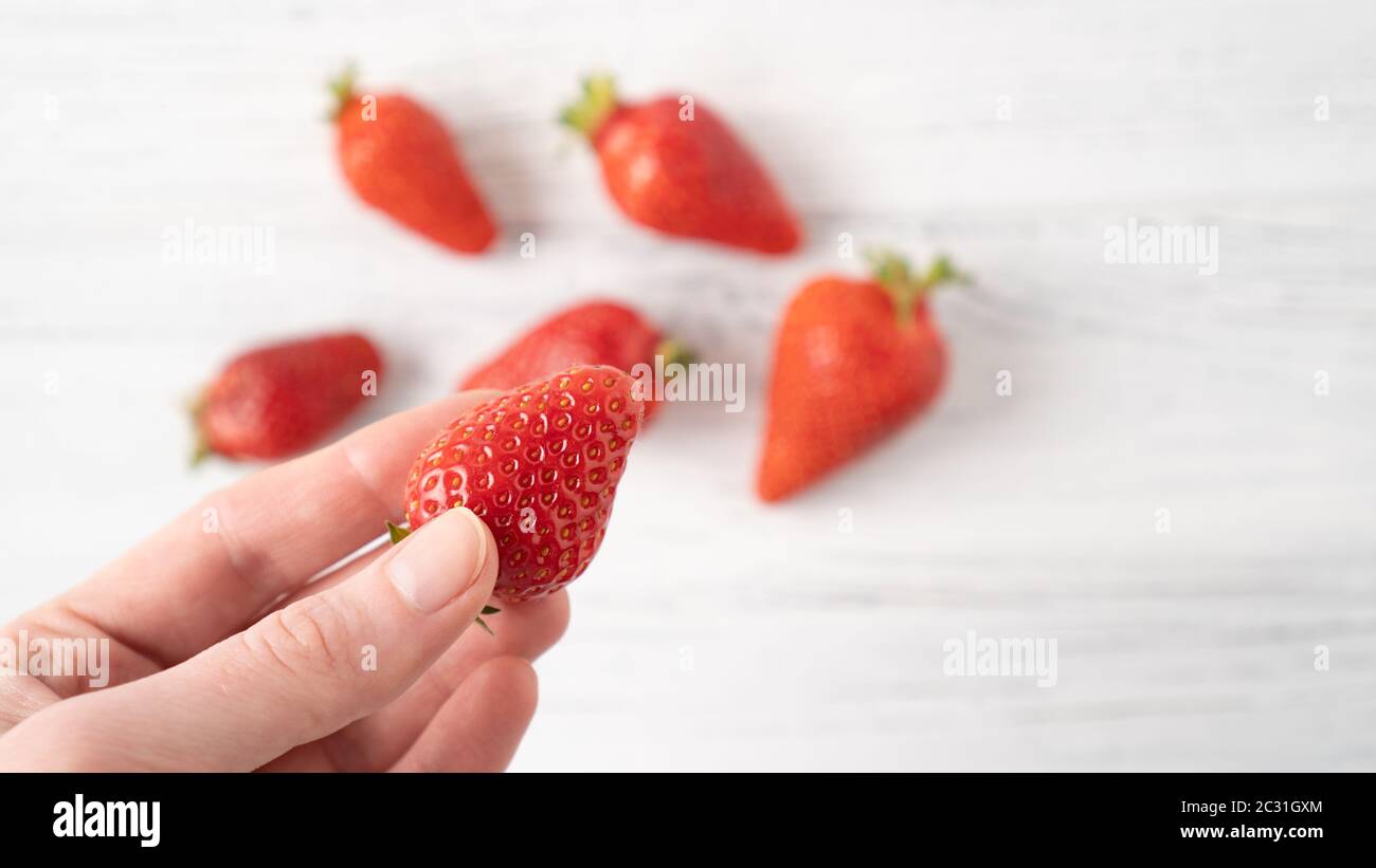 Hand holding ripe strawberry on white wooden background. Summer berry pleasure. Scattered strawberries Stock Photo