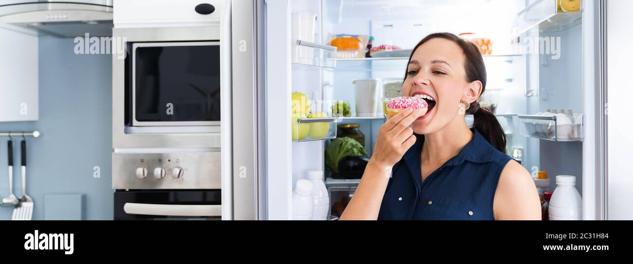Woman On Diet Indulging In Kitchen Near Refrigerator Stock Photo