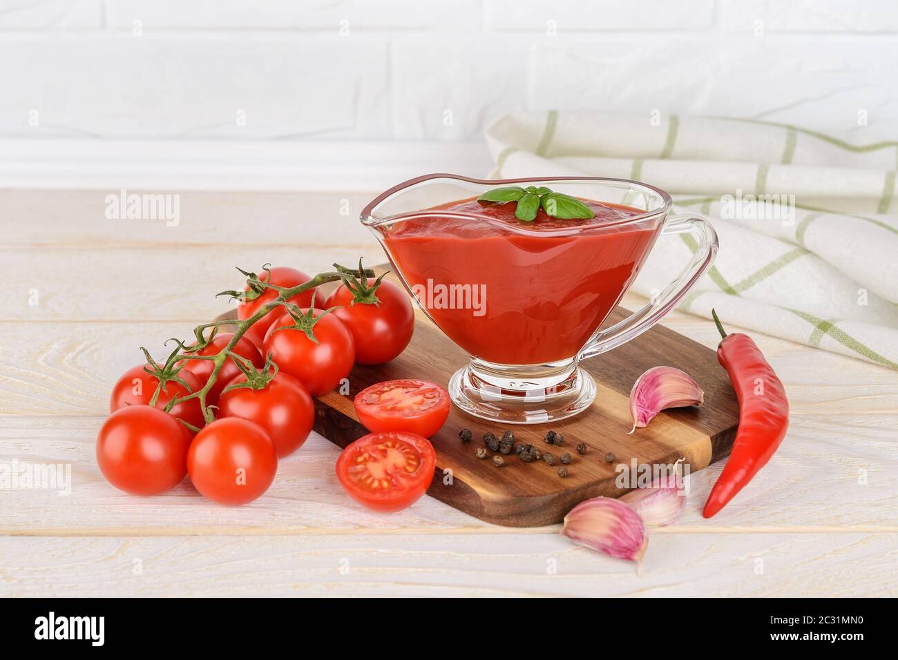 Thick tomato ketchup in a glass gravy boat and ripe cherry tomatoes, garlic and hot pepper on a wood kitchen table. Spices, seasonings and sauces. Stock Photo