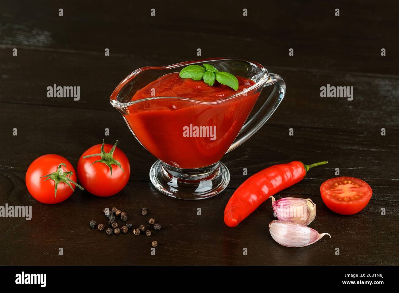 Tasty thick tomato ketchup in a glass gravy boat and red ripe cherry tomatoes, garlic and hot pepper on a wood table. Spices, seasonings and sauces. Stock Photo