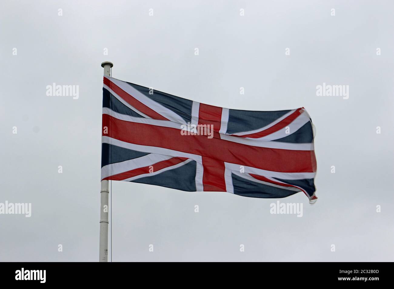 Union Jack flag fluttering in the wind on a white flagpole with a grey sky background. Stock Photo