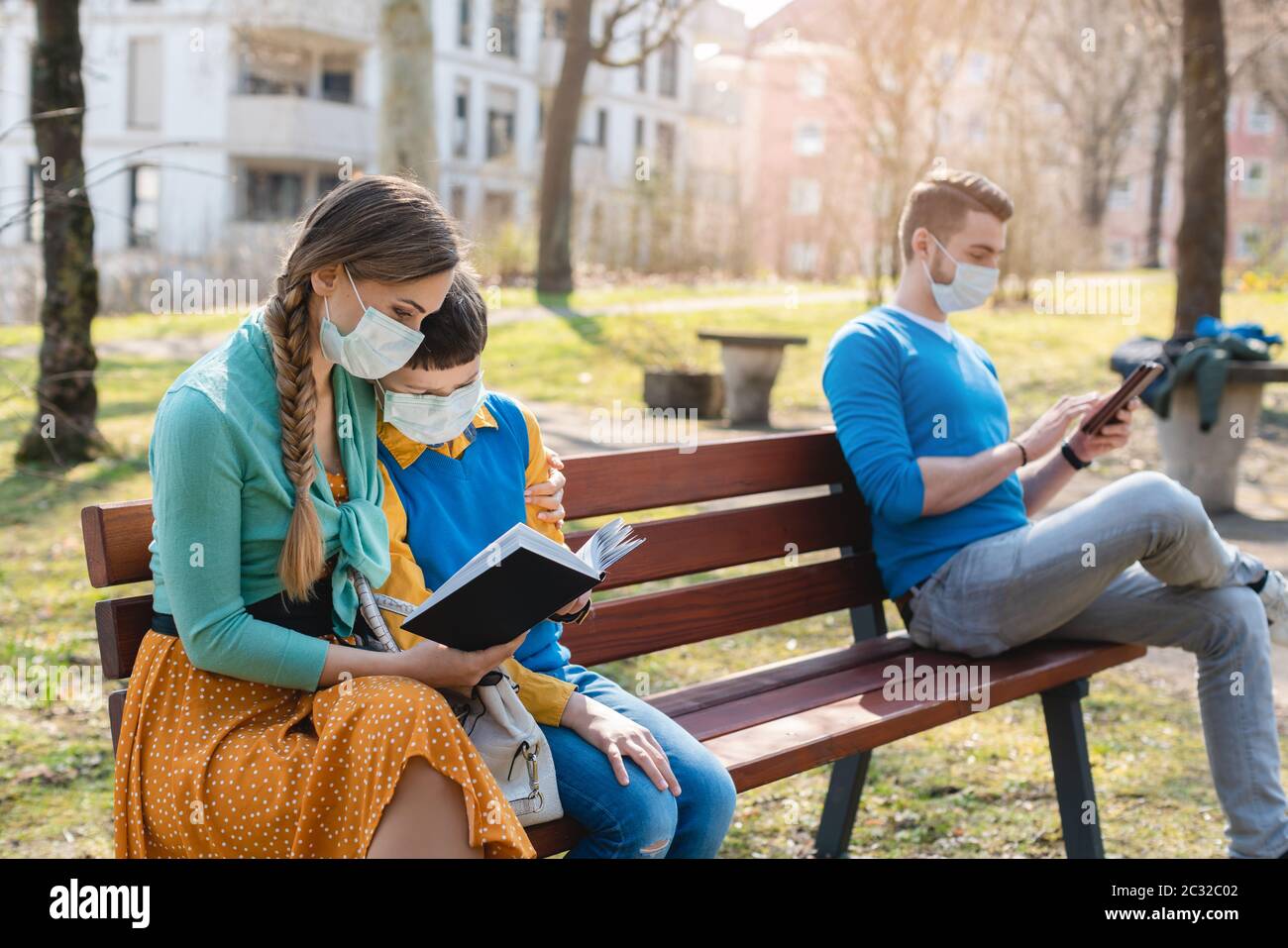 People sitting on park bench in the sun practicing social distancing in corona crisis wearing masks Stock Photo