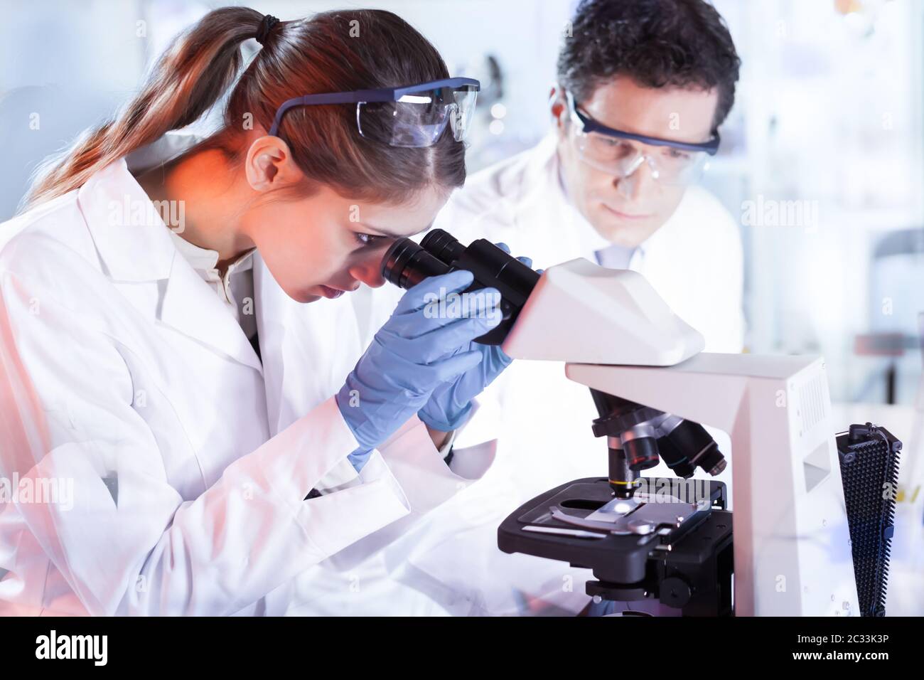 Life scientists researching in laboratory. Attractive female young scientist and her doctoral supervisor microscoping in their working environment. He Stock Photo