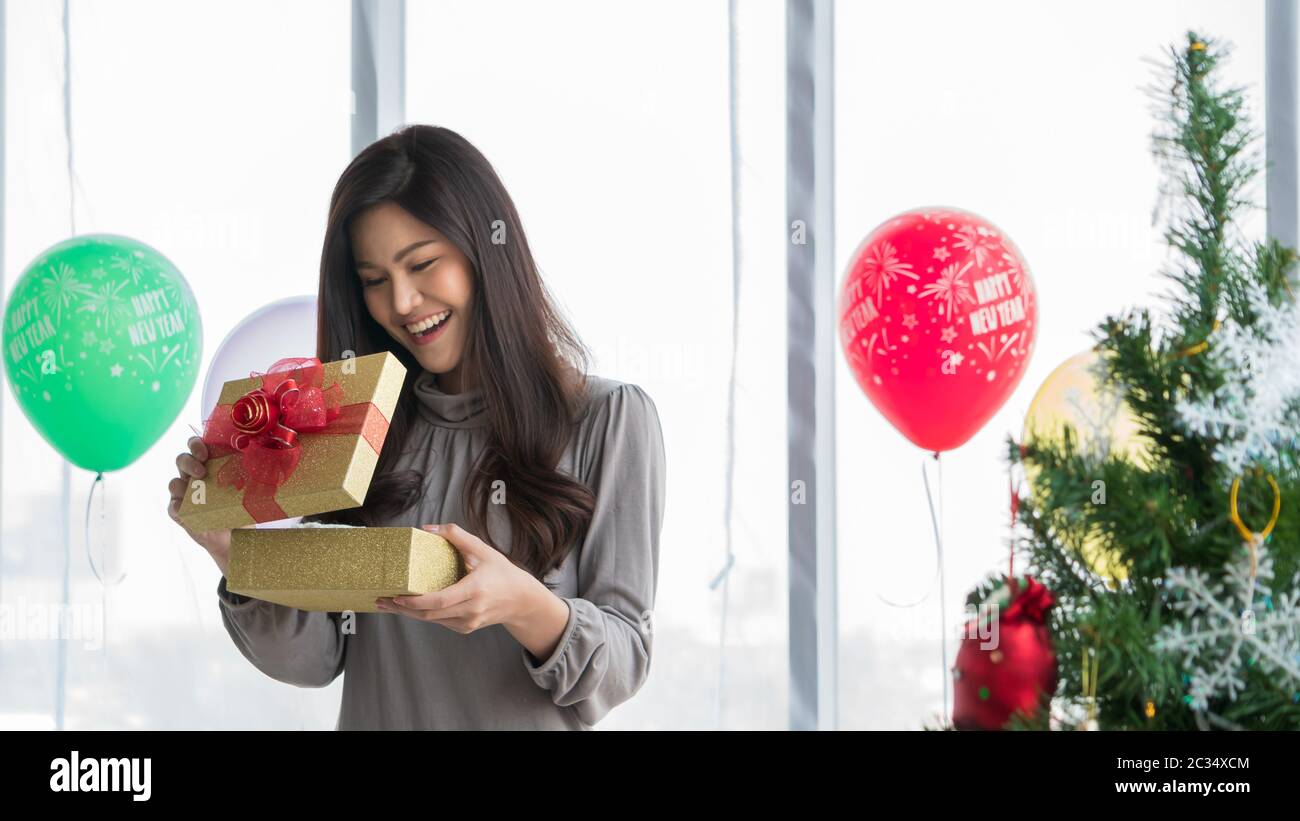 A beautiful Asian woman aged 20-30 years is smiling brightly and happily in a white room in her hand holding a golden gift box tied with a red bow as Stock Photo
