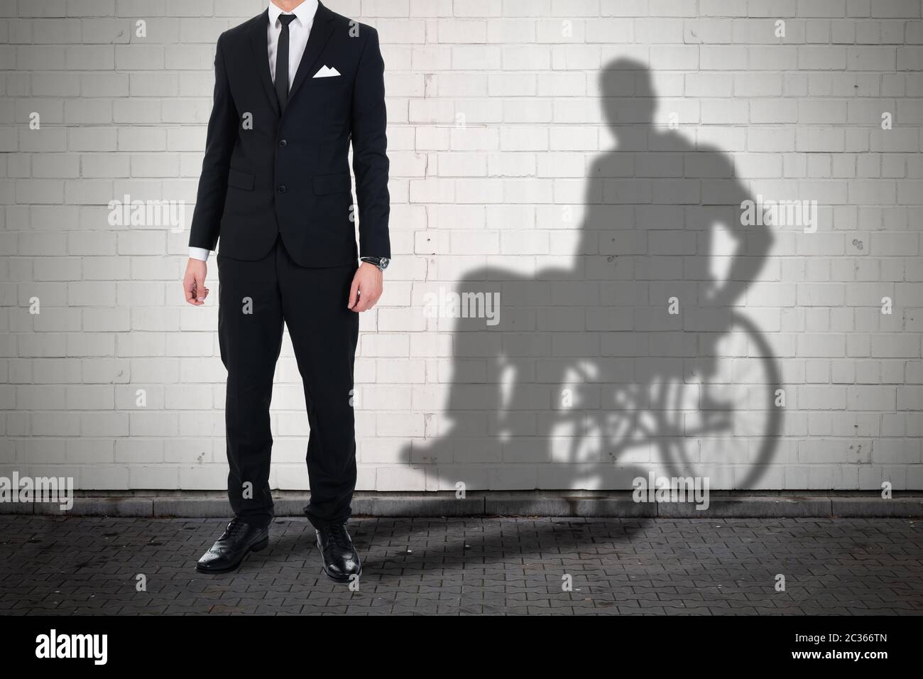 Businessman Standing Against White Brick Wall With Shadow Of A Disable Man Sitting On Wheelchair Stock Photo