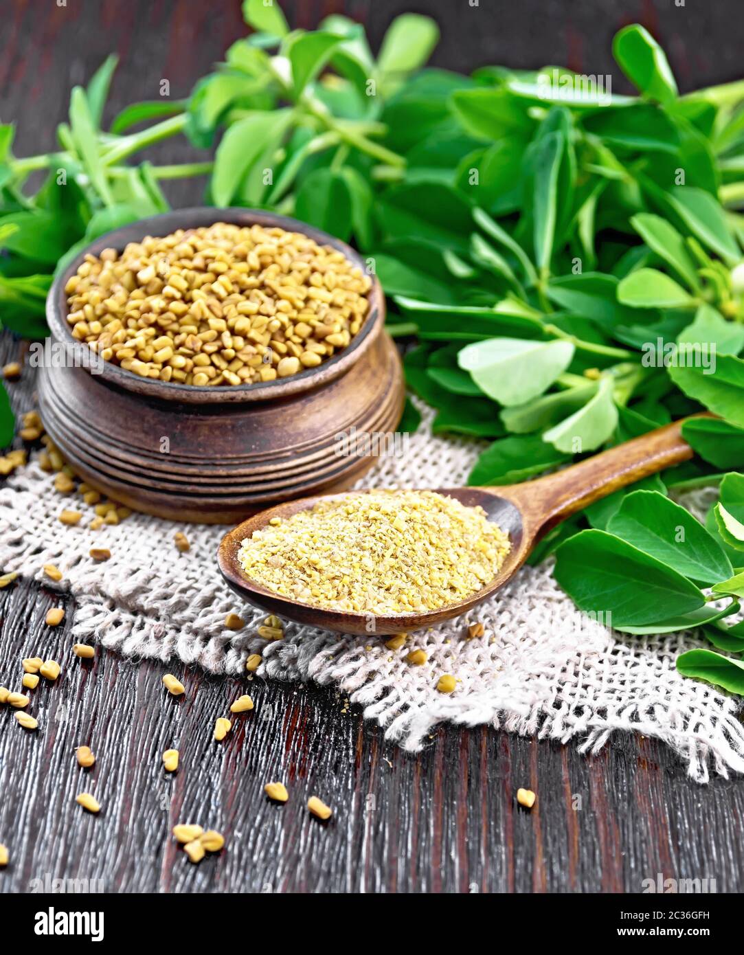 Ground fenugreek in a spoon and seeds in a bowl on burlap with green leaves on wooden board background Stock Photo