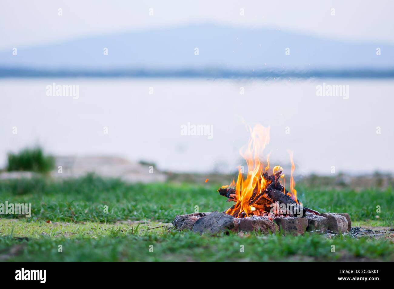 Small campfire with flames beside a lake with wood pile on the side. Stock Photo