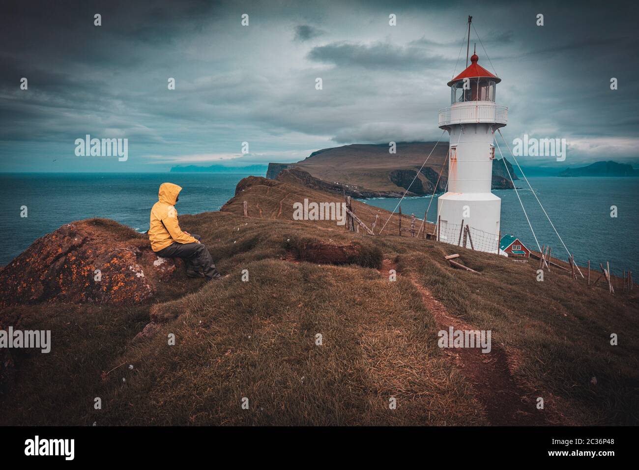 lighthouse on the island of mykines in the faroe, man with a yellow rain jacket sitting on the grass. Stock Photo