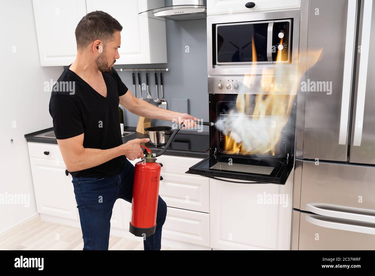 Man Using Fire Extinguisher To Put Out Fire From Oven At Home Stock