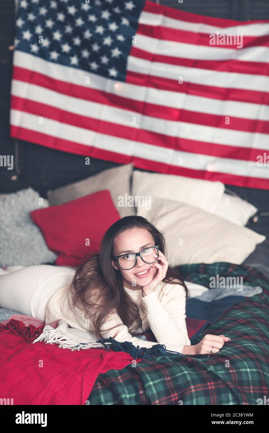 Pretty female in casualwear on bed with American flag on the wall Stock Photo