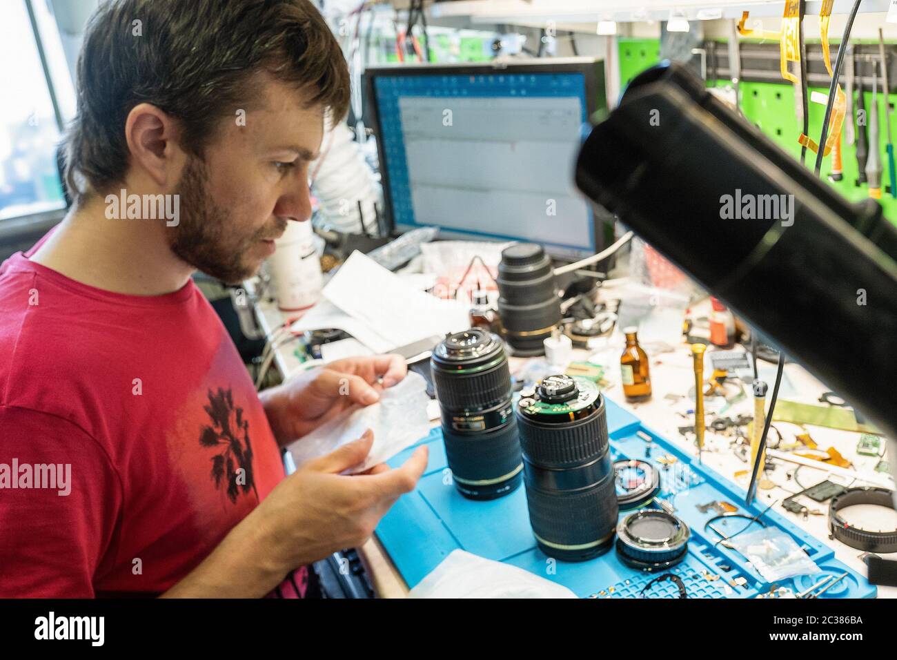 Master checks photo lenses in repair shop. Bearded man in red t-shirt works at workplace with master's tools on it. Service, repair and maintenance of Stock Photo