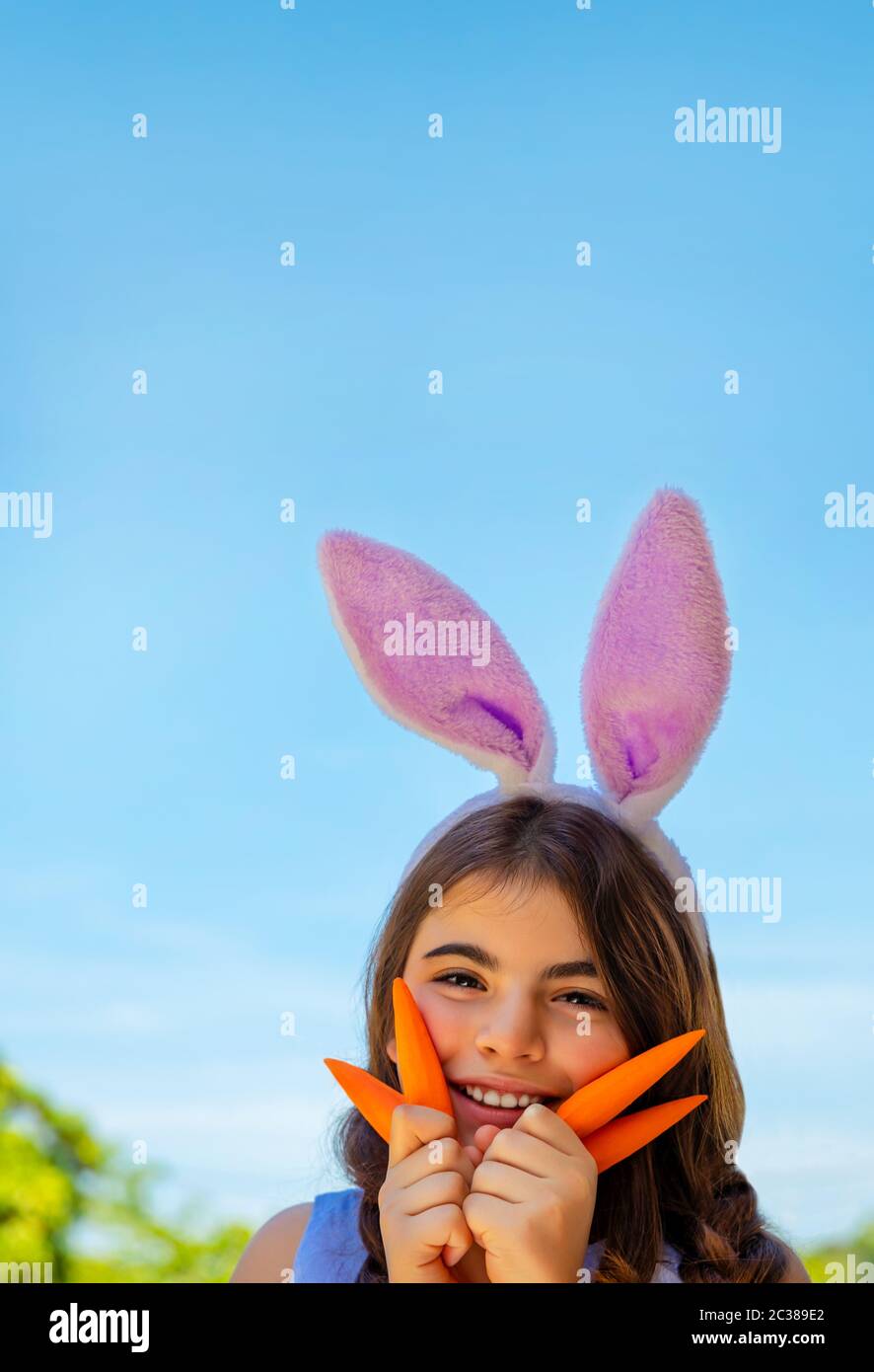 Portrait of a cute little bunny girl with carrots enjoying Easter, having fun gardening in the yard on a happy sunny day, traditional outfit for sprin Stock Photo
