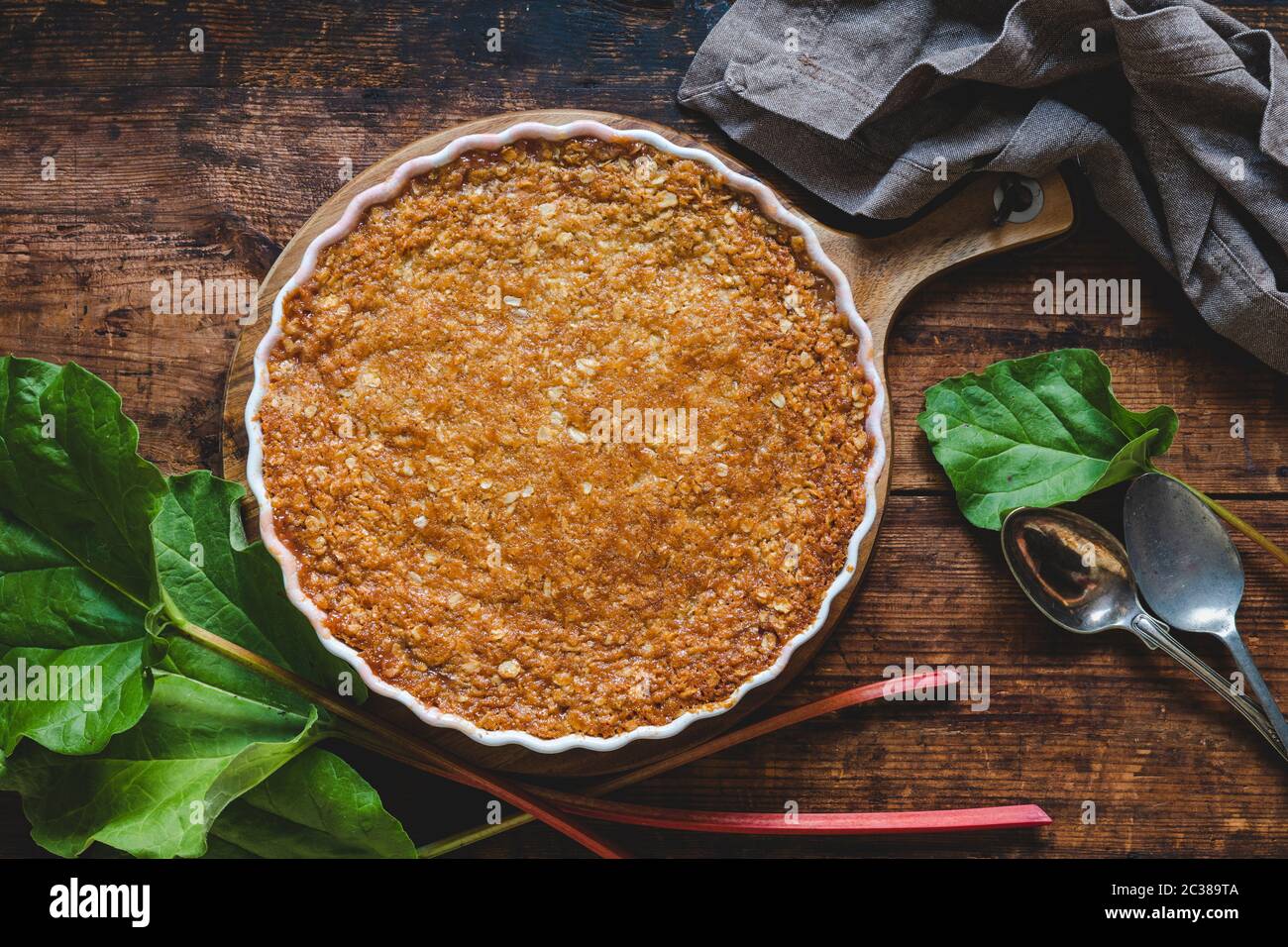 Traditional rhubarb pie on a rustic wooden table. The pie is with a toffee topping made of rolled oats, cream, syrup, sugar, flour and butter. Some sw Stock Photo