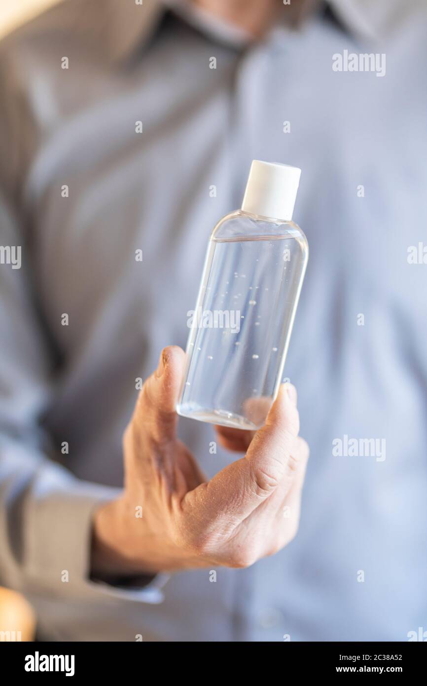 A male hand holding a small seethrough bottle containing sanitizer gel. The man is defocused in the background and he is  wearing a blue business shir Stock Photo