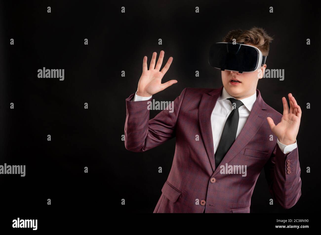 Law student with blond hair dressed in burgundy jacket, white shirt and black tie wearing virtual reality goggles posing on isolated black background Stock Photo