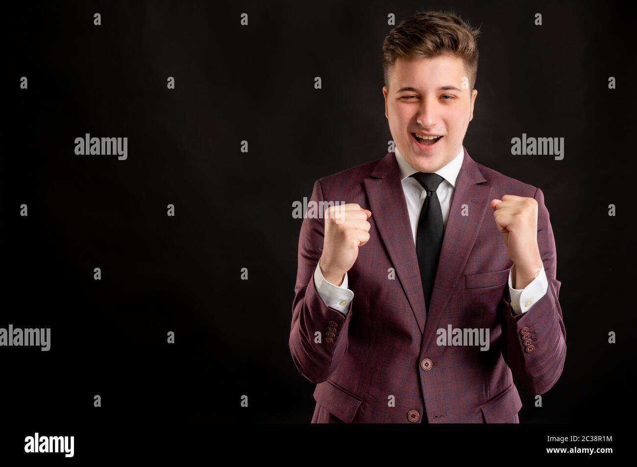 Law student with blond hair dressed in burgundy jacket, white shirt and black tie raised fists up exclaiming with joy and excitement posing on isolate Stock Photo