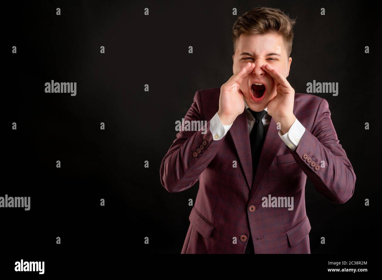 Law student with blond hair dressed in burgundy jacket, white shirt and black tie shouting with his hands to his mouth posing on isolated black backgr Stock Photo
