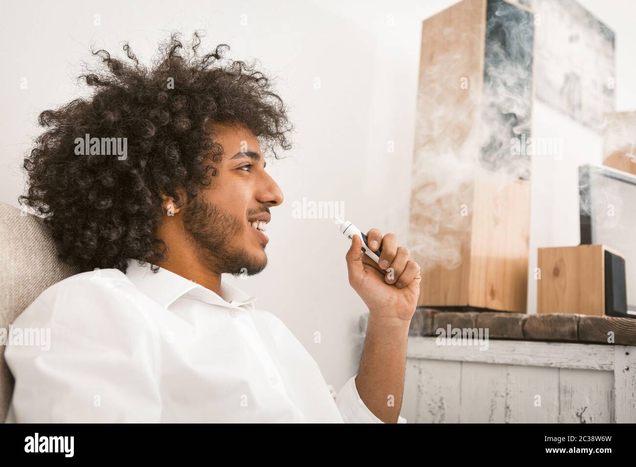 Shaggy man rests smoking e-cigarette. Smiling sun-tanned guy admires looking at smoke clouds. Alternative smoking concept. Side view. Close up shot Stock Photo