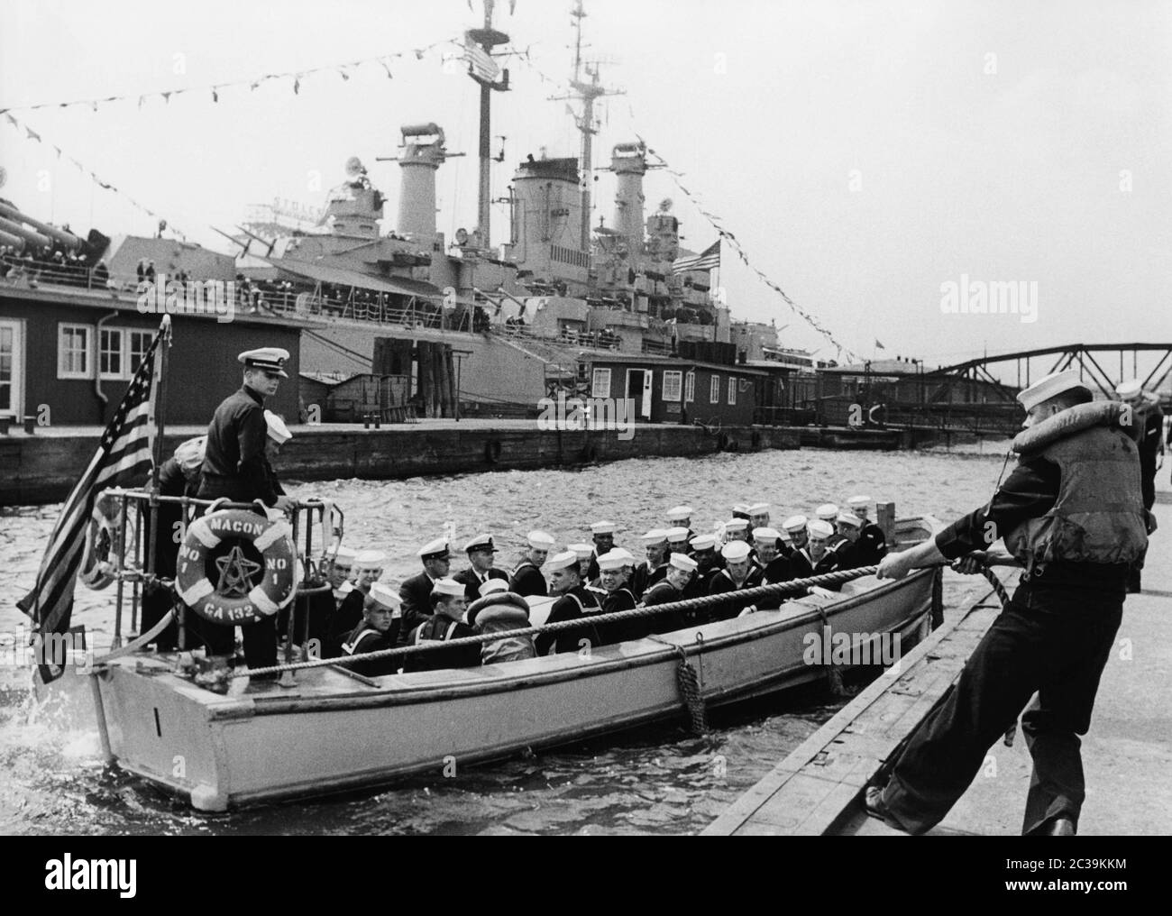 A dinghy of the heavy cruiser USS Macon (CA-132, seen in the background), is just mooring at the jetty. Sailors and officers of the crew are sitting in the boat. The occasion is a visit of the US Navy in Hamburg in the 1950s. Stock Photo