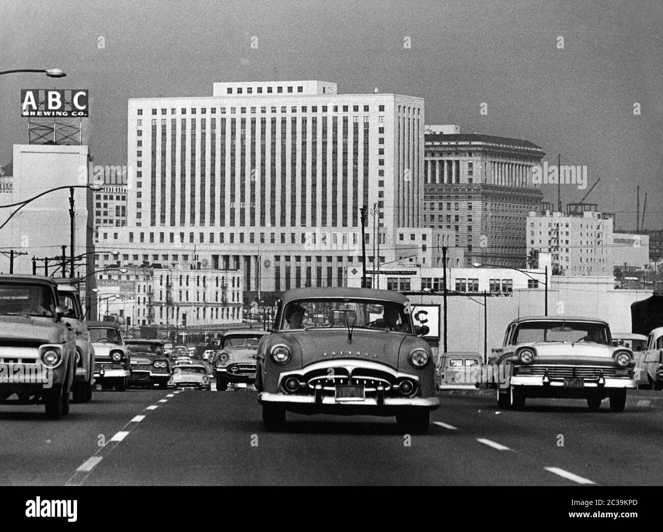 Dense traffic on the Santa Ana Freeway in Los Angeles in the 1960s with the typical car bodies of the 50s. On the upper left side there is a sign of the ABC Brewing Company in front of the skyscrapers of the city center. Behind, in the middle of the picture is the Federal Courthouse Building (seat of the Los Angeles Superior Court). On the right behind it is the Hall of Justice Building. Stock Photo