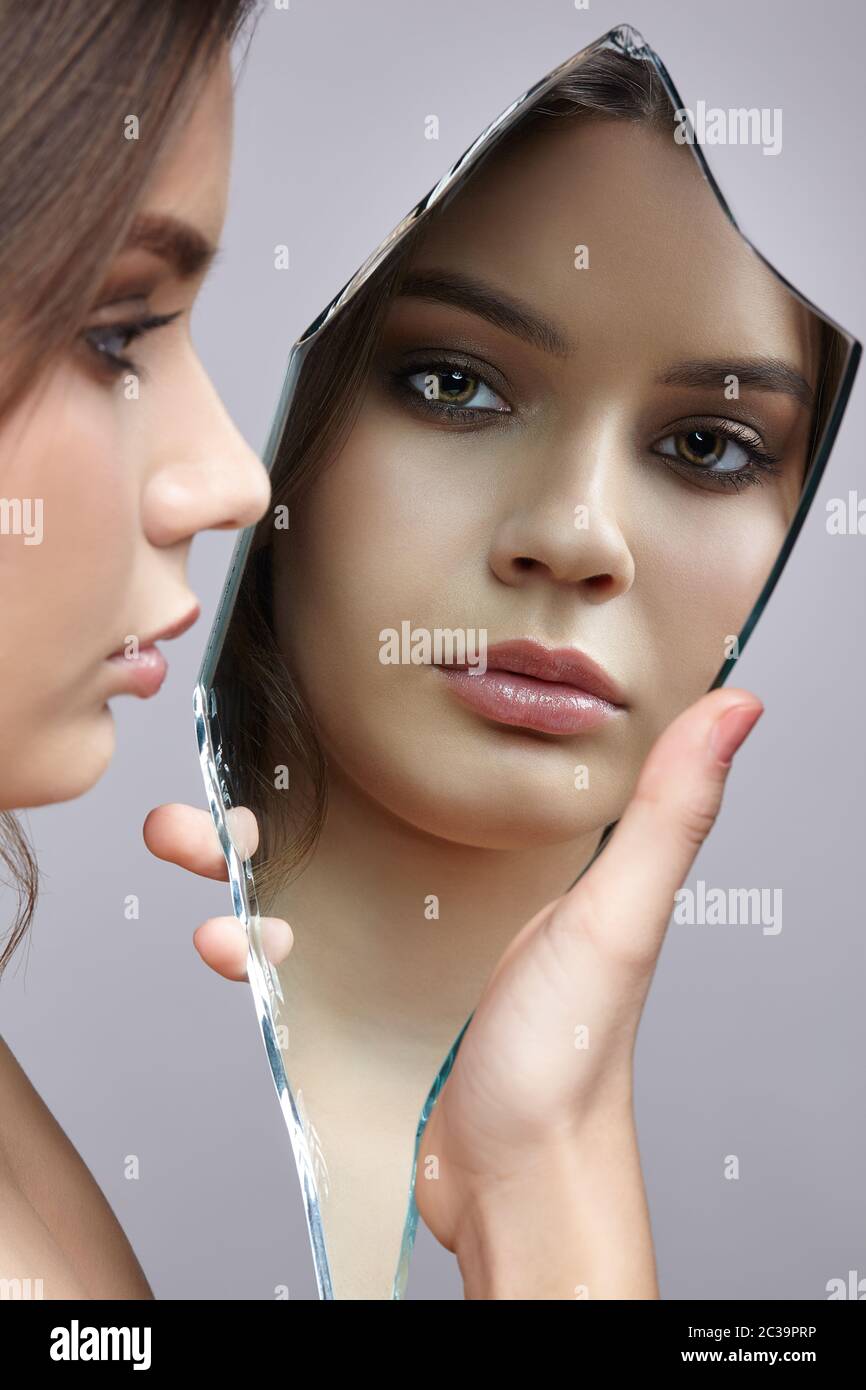 Female with mirror shard in hand posing on gray background. Face reflection in mirror splinter