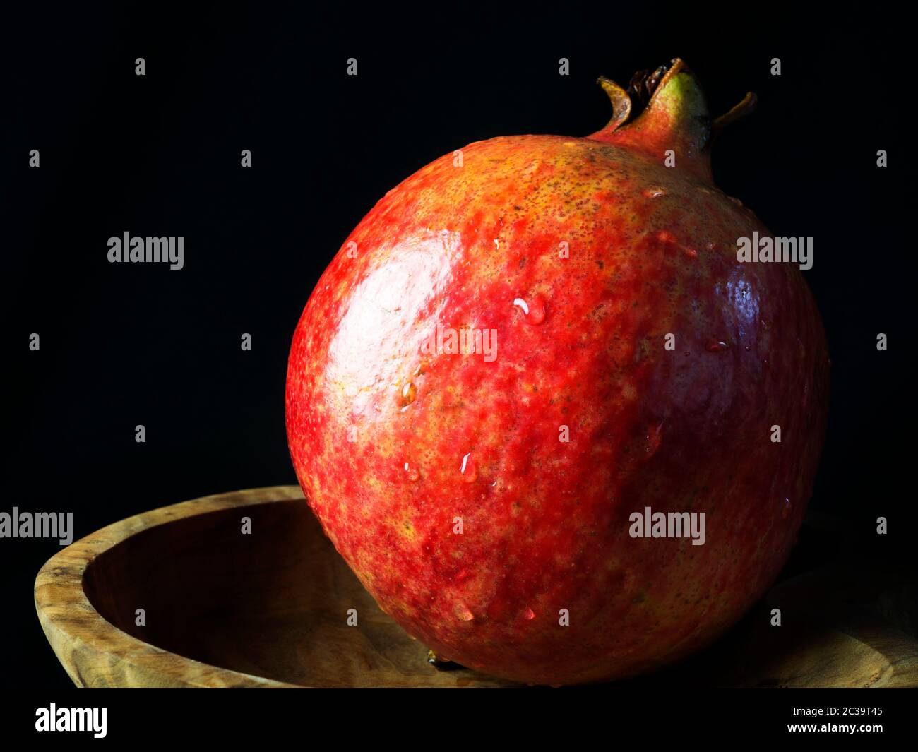 A single washed pomegranate with water droplets in a wooden dish against a black background Stock Photo