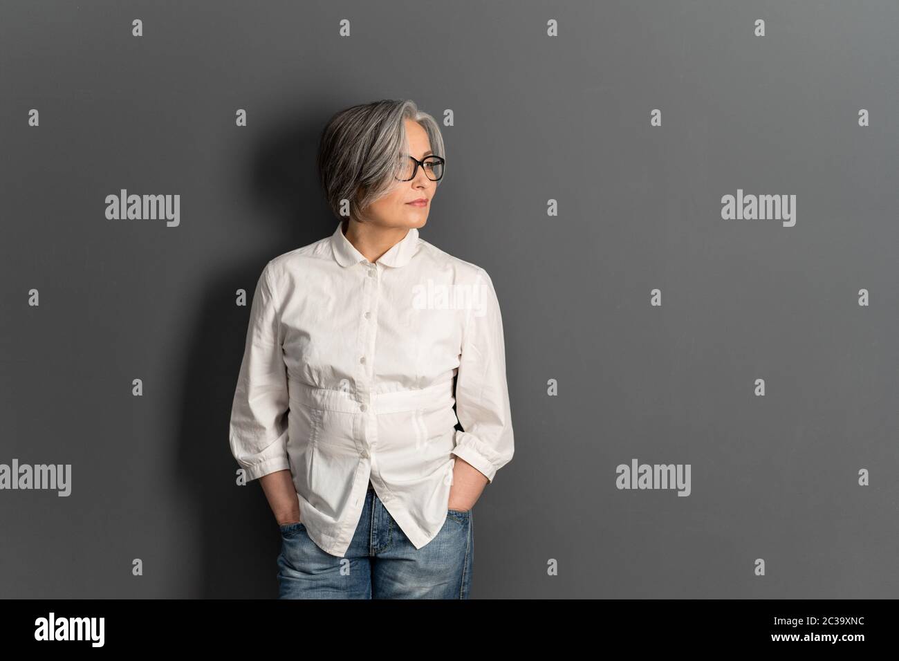 Beautiful mature lady in glasses stands hands in pockets looking at side. Elegant Gray-haired woman in whiite shirt on gray background. Copy space for Stock Photo