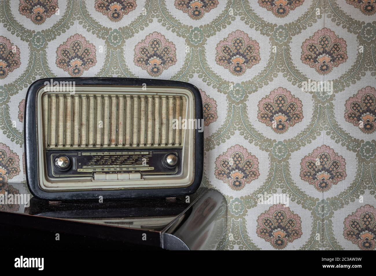 An old transistor radio, with knobs and buttons for manual tuning. In the background a vintage wallpaper. Ancient object, worn and ruined by time. Stock Photo