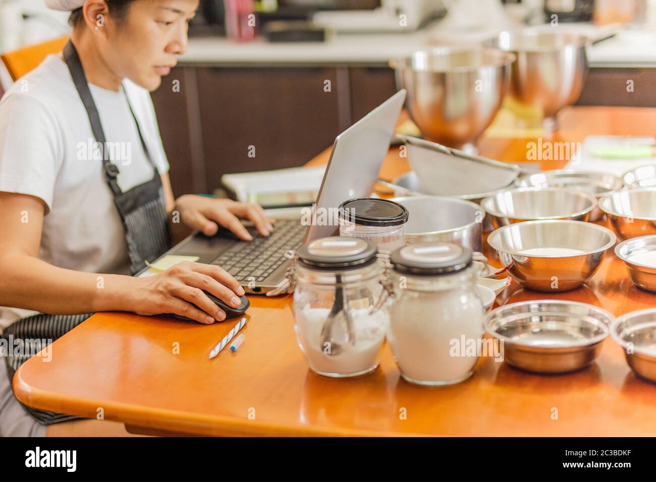 Female baker working on laptop with bakery ingredient on table. Stock Photo