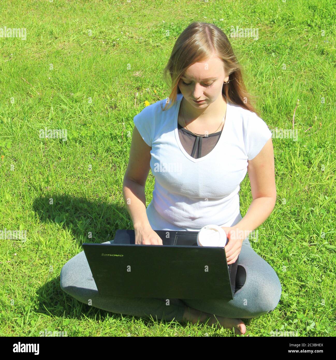 A beautiful young white girl in a white t-shirt and with long hair sitting on green grass, on the lawn and working behind a black laptop and holding a paper cup with coffee. Stock Photo
